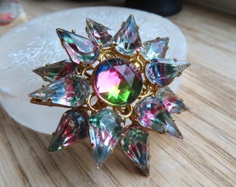 May include: A gold-tone brooch featuring a large, round, faceted center stone with iridescent pink and green hues, surrounded by a starburst arrangement of twelve pointed, pear-shaped, iridescent crystals set in a metallic gold-tone frame.