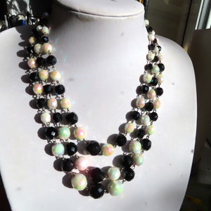 May include: A three-strand necklace featuring black, white, and iridescent beads. The beads are round and faceted, connected by a silver-coloured chain. The necklace is displayed on a white mannequin bust.
