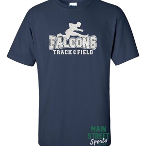 Op de afbeelding: Donkerblauw T-shirt met een wit silhouet van een hardloper en de tekst "Falcons Track & Field". De rechteronderhoek van het shirt heeft de tekst "Main Street Sports" in groen.