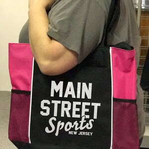 Puede incluir: Bolso tote negro con paneles laterales rosas y bolsillos de malla. El bolso presenta el texto "MAIN STREET Sports NEW JERSEY" en blanco. Tiene una correa negra para el hombro y ribete blanco.