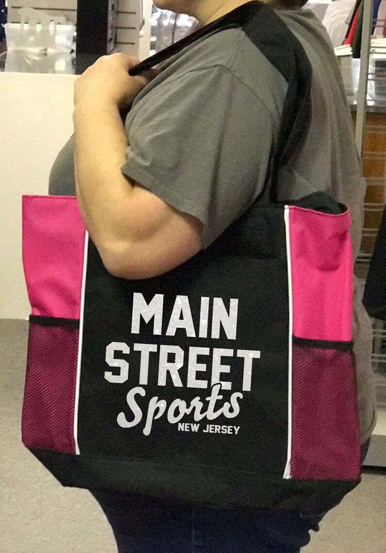 Puede incluir: Bolso tote negro con paneles laterales rosas y bolsillos de malla. El bolso presenta el texto "MAIN STREET Sports NEW JERSEY" en blanco. Tiene una correa negra para el hombro y ribete blanco.
