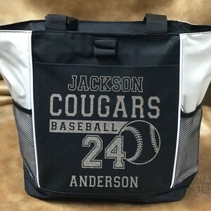 Puede incluir: Bolso de mano de b&eacute;isbol en blanco y negro con el texto "Jackson Cougars Baseball 24 Anderson" y un gr&aacute;fico de b&eacute;isbol. La bolsa tiene dos bolsillos laterales de malla y dos asas negras.
