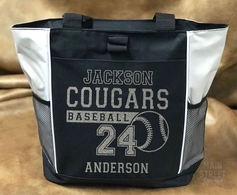 Puede incluir: Bolso de mano de b&eacute;isbol en blanco y negro con el texto "Jackson Cougars Baseball 24 Anderson" y un gr&aacute;fico de b&eacute;isbol. La bolsa tiene dos bolsillos laterales de malla y dos asas negras.