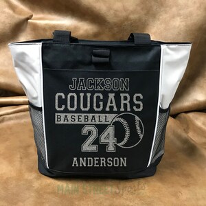 Puede incluir: Bolso tote negro y blanco con el texto "JACKSON COUGARS BASEBALL 24 ANDERSON" y un gr&aacute;fico de b&eacute;isbol. La bolsa tiene dos bolsillos laterales de malla y detalles en blanco.