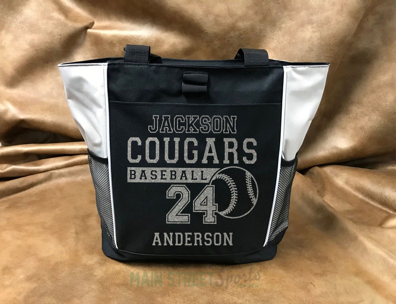 Puede incluir: Bolso tote negro y blanco con el texto "JACKSON COUGARS BASEBALL 24 ANDERSON" y un gr&aacute;fico de b&eacute;isbol. La bolsa tiene dos bolsillos laterales de malla y detalles en blanco.