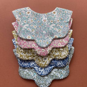 May include: A stack of five scalloped-edge bibs featuring various floral patterns. The top bib has a light blue background with white and pink flowers. Below it are bibs in pink, olive green, navy blue, and sage green, all with floral prints.
