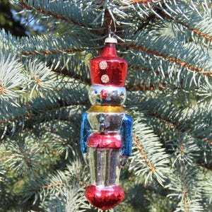 May include: A vintage Christmas bauble in the shape of a toy soldier. The ornament has a red hat, blue jacket, and silver body. It hangs from a silver hook against blue-green pine needles. A festive holiday decoration.