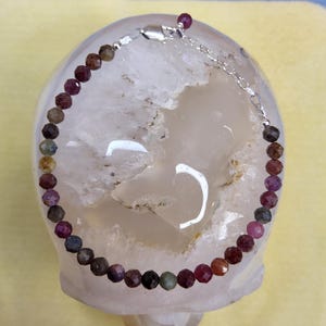 May include: A gemstone bracelet featuring faceted, multi-colored beads in shades of red, brown, green, and gray. The bracelet is strung on a silver chain with a lobster clasp. The beads are approximately 0.2 inches in diameter.