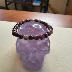 May include: A purple and green gemstone bracelet on a purple crystal skull. The bracelet is made of round beads.