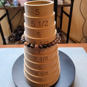 May include: A wooden ring sizer with markings for sizes 5, 5 1/2, 6, 6 1/2, 7, 7 1/2, 8, 8 1/2, 9, and 9 1/2. A brown and gold beaded bracelet is resting on the sizer.