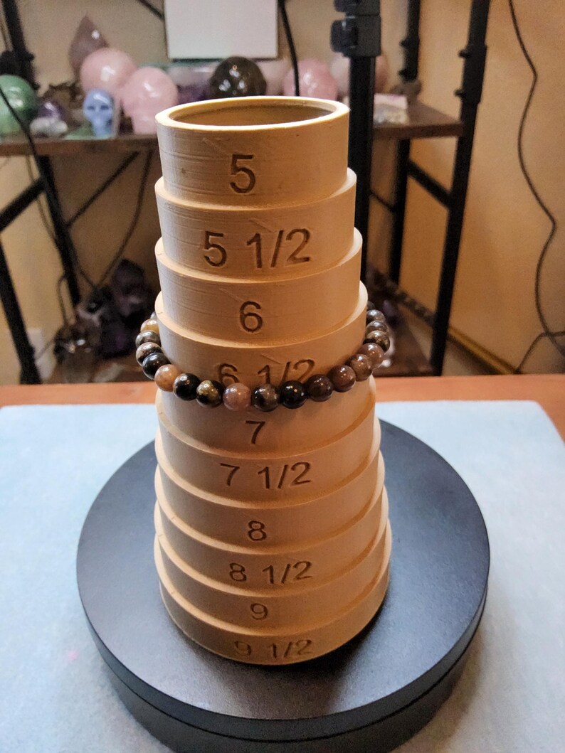 May include: A wooden ring sizer with markings for sizes 5, 5 1/2, 6, 6 1/2, 7, 7 1/2, 8, 8 1/2, 9, and 9 1/2. A brown and gold beaded bracelet is resting on the sizer.