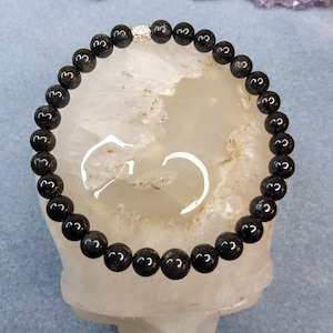 May include: A circular beaded bracelet composed of smooth, polished black spherical beads. The bracelet features a single, small, cylindrical silver-toned metal spacer bead with a textured, intricate pattern positioned at the top center.