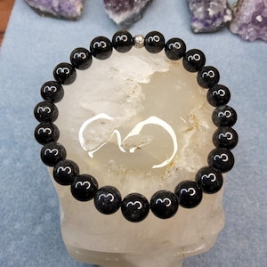 May include: A circular beaded bracelet composed of smooth, polished black spherical beads. The bracelet features a single small, textured silver-toned metal spacer bead positioned at the top center, creating a contrast against the dark stones.