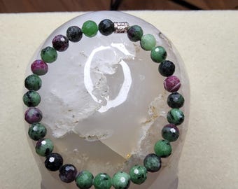 May include: A green, black, and pink gemstone bracelet with a silver clasp. The bracelet is made of round beads and is displayed on a white surface.