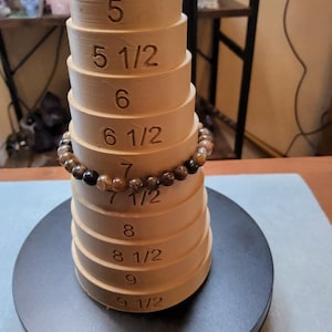May include: A set of wooden ring sizers, stacked in a cone shape, with a brown and black beaded bracelet around the middle. The sizers are labeled with numbers from 5 to 9 1/2.