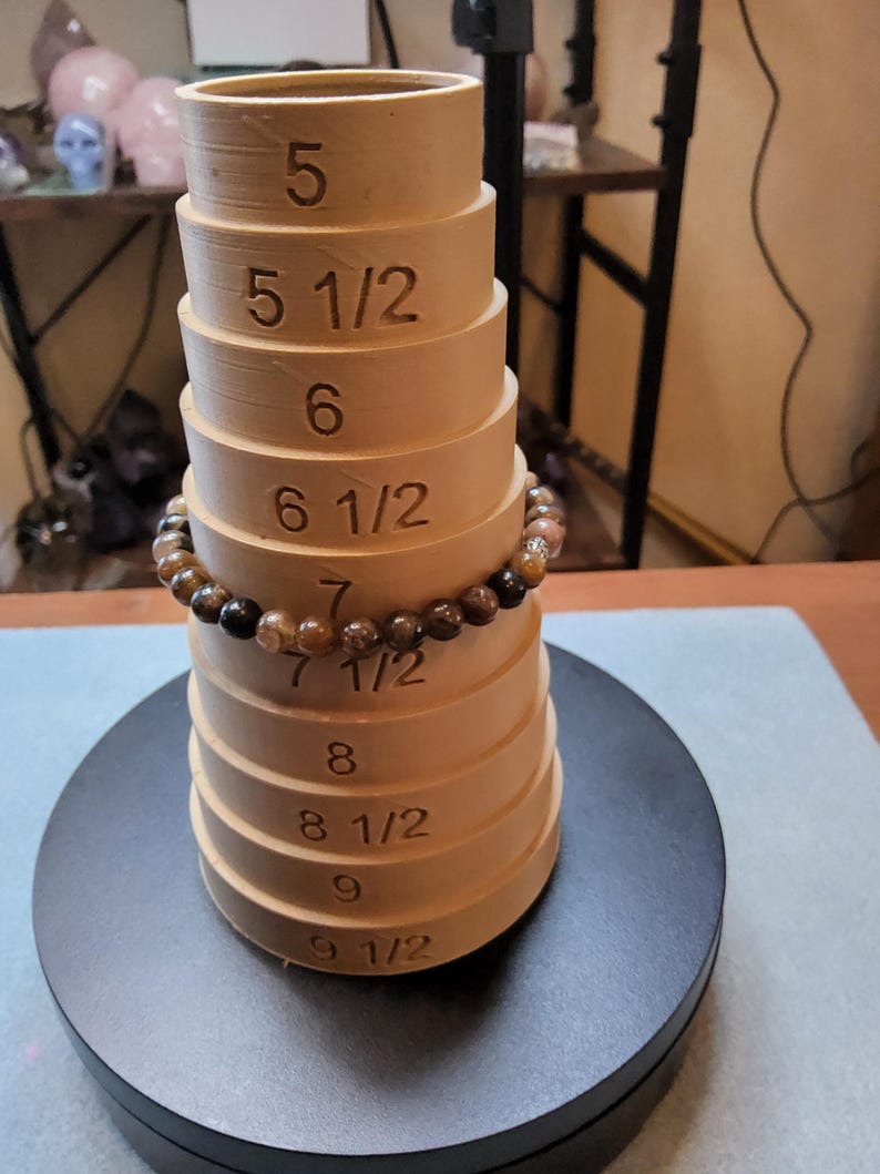 May include: A set of wooden ring sizers, stacked in a cone shape, with a brown and black beaded bracelet around the middle. The sizers are labeled with numbers from 5 to 9 1/2.