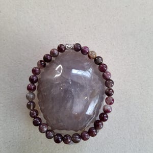 Puede incluir: Un brazalete de piedras preciosas púrpura y verde con un cierre plateado. El brazalete está hecho de cuentas redondas y está sobre una superficie blanca.