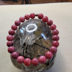 May include: A pink gemstone bracelet with a silver clasp. The bracelet is made of round beads and is lying on a clear crystal.
