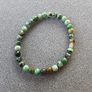 May include: A green and brown gemstone bracelet with a gold accent. The bracelet is made of round beads and is strung on a clear elastic cord.