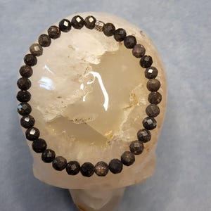May include: A dark gray beaded bracelet with faceted beads and a small silver accent bead. The bracelet is resting on a white, rounded surface against a light blue background. The beads appear to be approximately 0.3 inches in diameter.