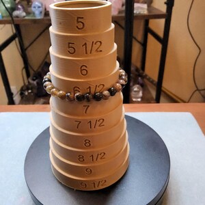May include: A wooden ring sizer with a brown finish. The sizer has a series of graduated rings with numbers indicating sizes from 5 to 9 1/2. A brown gemstone bracelet is resting on the sizer.