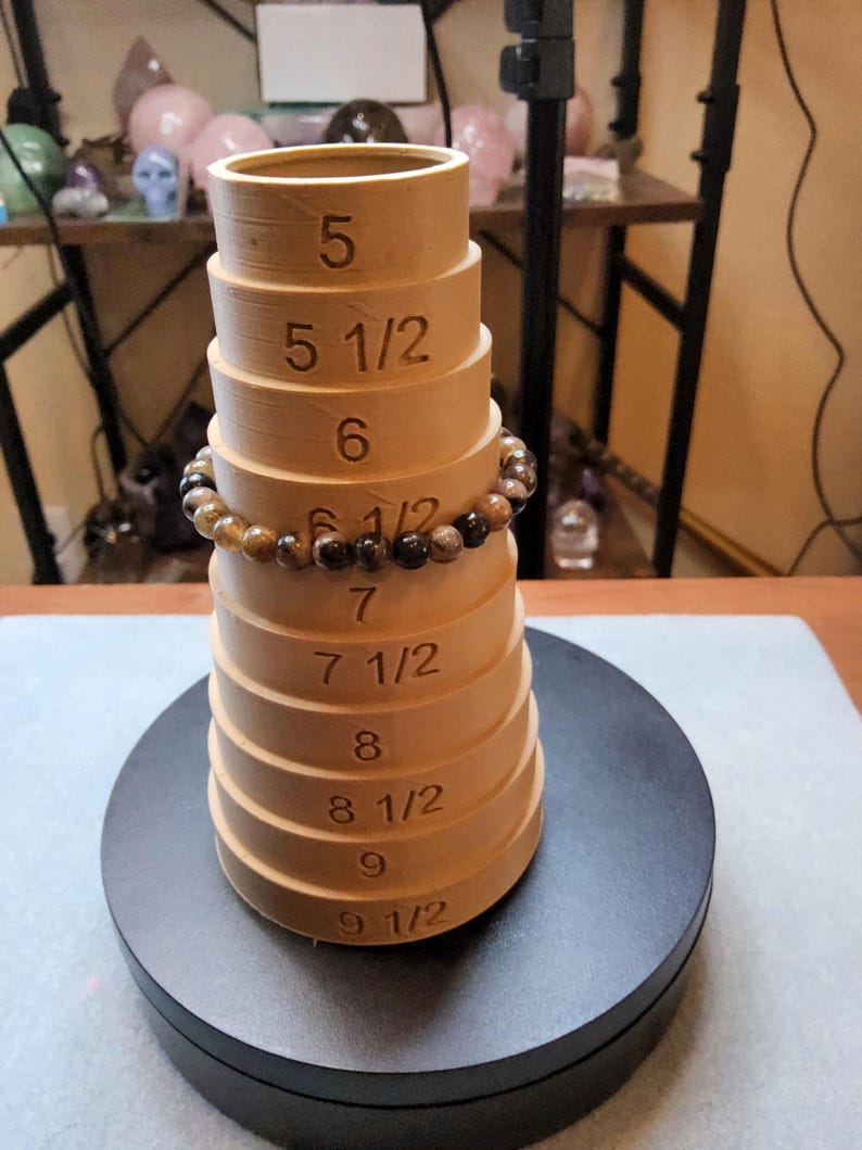 May include: A wooden ring sizer with a brown finish. The sizer has a series of graduated rings with numbers indicating sizes from 5 to 9 1/2. A brown gemstone bracelet is resting on the sizer.