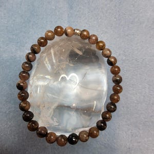 May include: A brown and black gemstone bracelet with a silver bead accent. The bracelet is made of round beads and is displayed on a clear crystal.