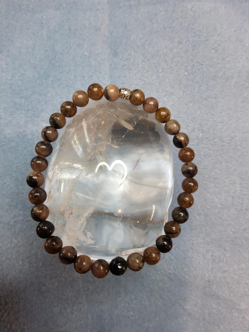 May include: A brown and black gemstone bracelet with a silver bead accent. The bracelet is made of round beads and is displayed on a clear crystal.