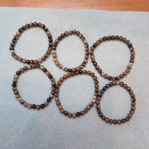 May include: Six brown and black beaded bracelets with silver accents. The bracelets are arranged in a circle on a light blue surface.