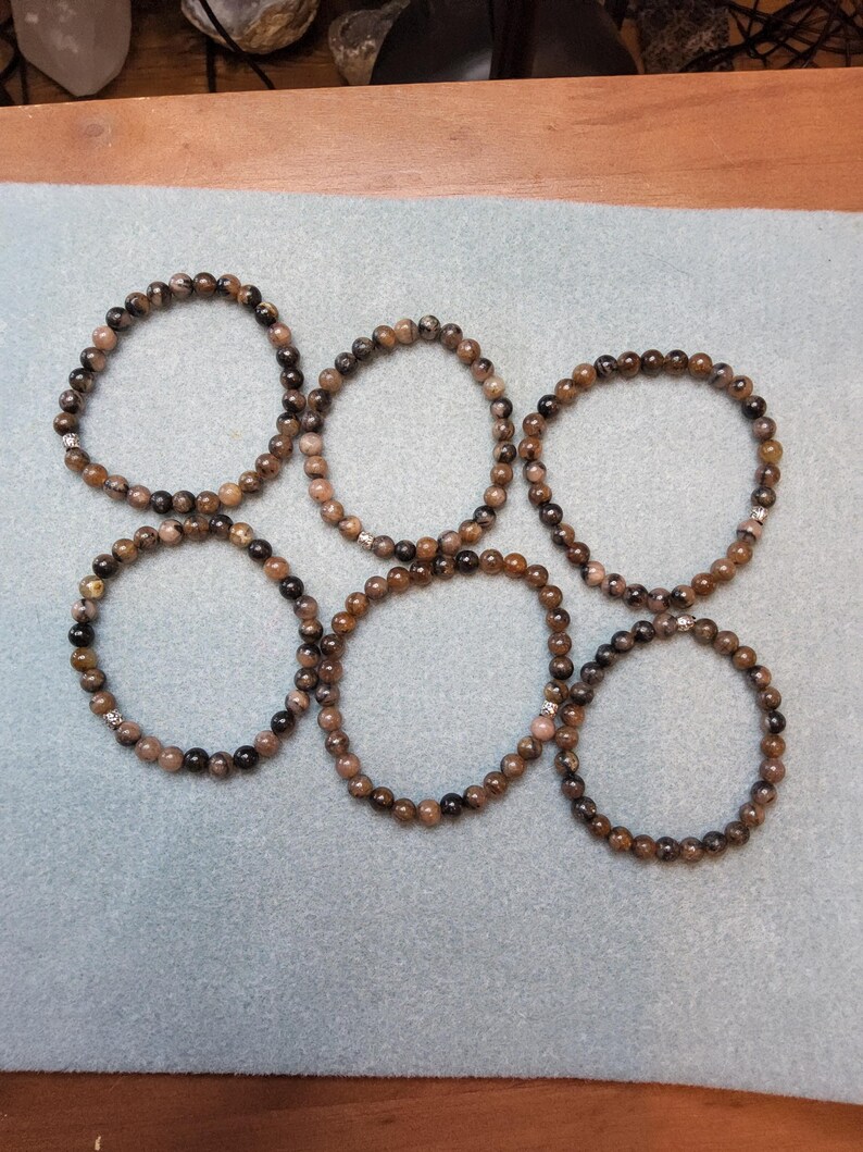 May include: Six brown and black beaded bracelets with silver accents. The bracelets are arranged in a circle on a light blue surface.