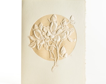 May include: A white card with a raised, embossed design of a flower in a circle. The flower is white and the circle is a light brown color.