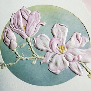 Könnte beinhalten: Eine strukturierte, geprägte Grußkarte mit einer erhabenen, hellrosa Magnolienblüte und einer geschlossenen Knospe an einem Zweig. Das florale Design ist vor einem kreisförmigen, gedämpften salbeigrünen Hintergrund platziert und wirkt dreidimensional.