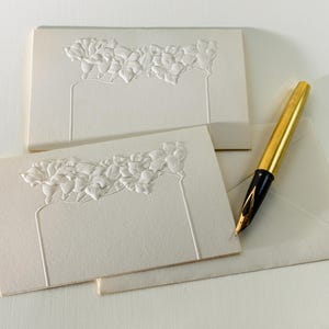 May include: Two white blank note cards with an embossed floral design. A gold pen with a black nib is lying on top of the cards.