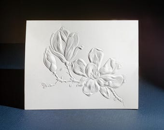 May include: A rectangular white greeting card featuring a central embossed design of a magnolia branch with one fully bloomed flower, one partially open flower, and one closed bud, all rendered in a raised, monochromatic white texture.