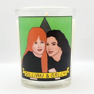 May include: A candle with a green background and a yellow banner that says "Gillian & Sally". The candle features a cartoon illustration of two women with red and black hair.