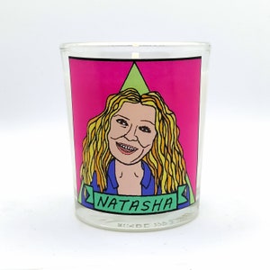 May include: A clear glass candle holder with a pink background featuring a cartoon illustration of a woman with blonde hair. The woman's name, "Natasha," is displayed on a green banner below her image. The candle holder is filled with a white wax.