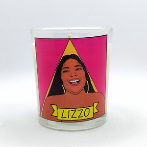 Puede incluir: Una vela de vidrio transparente con un gráfico de Lizzo sobre un fondo rosa con un triángulo amarillo. La palabra "Lizzo" está impresa en una pancarta amarilla. La vela está llena de cera blanca.