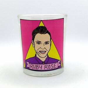 Puede incluir: Un portavelas de cristal transparente con un gráfico rosa y amarillo que presenta un retrato de Ruby Rose. El retrato está rodeado por un fondo rosa y un triángulo amarillo. El nombre "RUBY ROSE" está escrito en una pancarta morada.