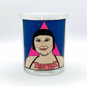 May include: A clear glass candle featuring a graphic of a woman with black hair and a pink triangle on a blue background. The name "BETH" is printed on a banner below the woman's image.