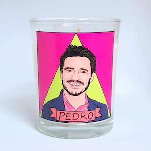 May include: A clear glass candle with a portrait of a man on the front. The portrait is set against a pink background with a green triangle. The man's name, "PEDRO", is printed on a banner below the portrait. The candle has a white wax and a single wick.