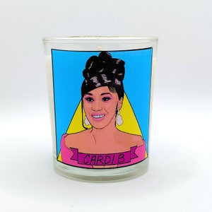 Puede incluir: Una vela de vidrio transparente con un gráfico de Cardi B sobre un fondo azul y amarillo. La vela tiene un interior de cera blanca y una mecha. El gráfico incluye el nombre "Cardi B" en una pancarta rosa.