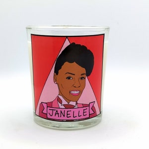 Puede incluir: Una vela de vidrio con un gráfico rojo y rosa de un retrato de mujer. El retrato está sobre un triángulo rojo, con el nombre "JANELLE" en una pancarta debajo. La vela tiene un interior de cera blanca.