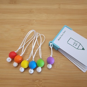 Peut inclure: Une collection de breloques de crayons perlés colorés avec des cordons blancs, disposées à côté d'une pile de cartes blanches maintenues ensemble par un classeur bleu. Les cartes portent le texte "Make Your Own" et une illustration de crayon souriant.