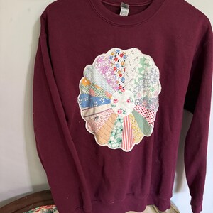 May include: A maroon sweatshirt with a large, colorful patchwork flower design on the front. The flower is made of different fabrics and patterns, including floral, plaid, and striped.