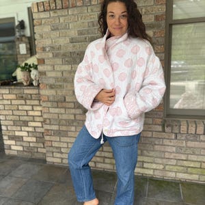 May include: A white quilted jacket with a pink seashell pattern. The jacket has a relaxed fit, long sleeves, and two front pockets. The person is wearing blue jeans.