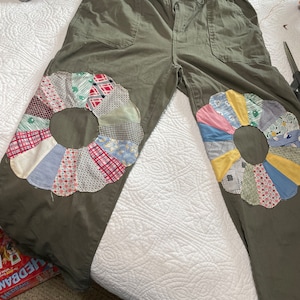 May include: Olive green cargo pants with a drawstring waist and patchwork flower designs on the knees. The flowers are made of various fabrics in shades of pink, blue, yellow, green, and red.