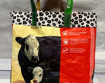 May include: A red, white, and green tote bag made from recycled feed bags. The bag features a black and white floral pattern and a picture of a cow and calf. The bag is labeled "CUBES" and has the text "E-Z OPEN" on the strap. The bag is designed to be easily fed on the ground and is a supplemental protein for all stages of breeding cows and bulls to support body condition. The bag is 50 lbs (22.67 kg).