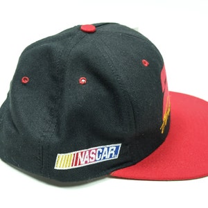 Peut inclure: Casquette de baseball noire et rouge avec un logo "NASCAR" jaune, bleu et rouge sur le devant. La casquette a une visi&egrave;re rouge.