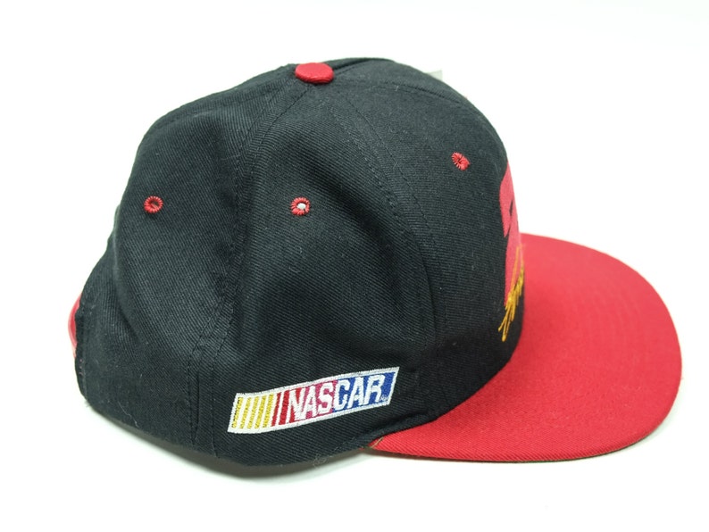 Peut inclure: Casquette de baseball noire et rouge avec un logo "NASCAR" jaune, bleu et rouge sur le devant. La casquette a une visi&egrave;re rouge.