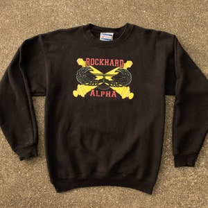 Peut inclure: Sweat-shirt noir avec un graphique jaune et rouge de canons croisés et d'ailes avec le texte "ROCKHARD ALPHA".